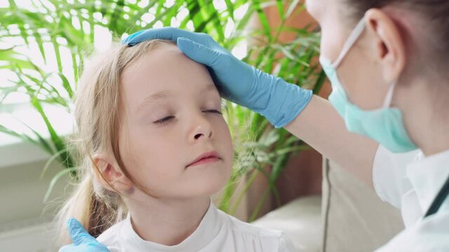 Young beautiful woman doctor examines a sick child at home, sitting on a sofa.