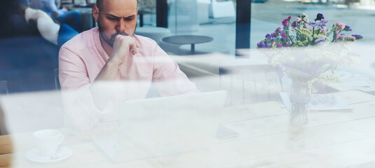 Fototapeta premium Puzzled and concerned male freelancer or student look to net-book, worried and thoughtful businessman or entrepreneur sitting front open laptop computer in modern coffee shop interior in urban setting
