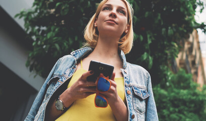 Low angle view with pretty young woman hold smartphone standing on mandarin tree background in city, lost female tourist using telephone for navigation, student girl browsing internet on mobile phone