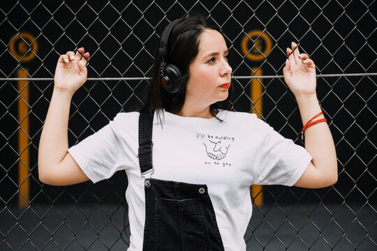 Joven mujer con overol y aud&iacute;fonos reposa en la reja de una cancha de squash