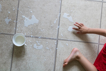Child's bare feet standing in puddle of spilled milk on floor with white cup.