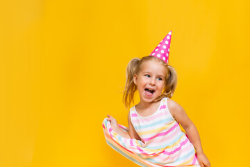 Happy birthday child girl with two ponytales in pink cap on colored yellow background shoing her...