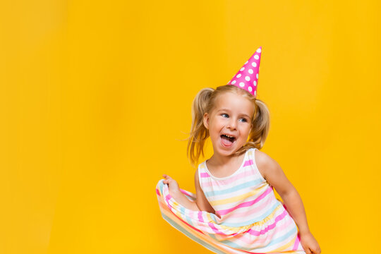 Happy birthday child girl with two ponytales in pink cap on colored yellow background shoing her tongue.
