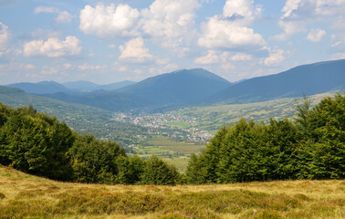 Beautiful nature mountain landscape with mountains, grassy fields, trees and Carpathian village in valley under blue sky with clouds in summer day