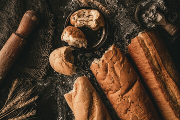 Broken bread baguettes on black slate background with wheat spikelets, sackcloth, flour. Pastries and bread in a bakery, Food concept, top view