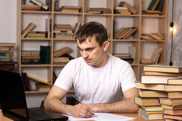 young male student preparing for the exam at home looks into a laptop on the background of a bookcase. businessman or office employee checks the work.