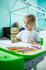 A girl in a white T-shirt sits in her room at the table and draws with colored pencils