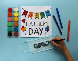 Child's hand draws a mustache on a protective mask as a gift for the dad. Greeting card Happy Father’s Day, paints and markers on violet background. Father’s day in the context of coronavirus pandemic