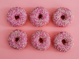 Six pink American donuts with white sprinkles lie in the middle on a pink paper background, close-up top view.