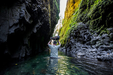 Adventurous man holding a backpack above his head while walking through a river in a gorge.