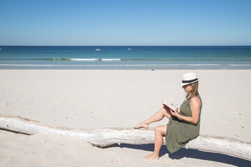 Middle-aged blonde woman sitting on a tree trunk in the beach reading a book