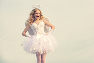 Innocent Girl with angel wings standing with bow and arrow against blue sky and white clouds. Portrait of a cupid little girl pray. Child with angelic character.