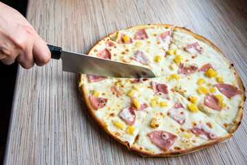 female hand cuts the pizza with a kitchen knife. Delicious pizza with ham, mozzarella and pineapple on wood table