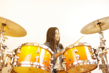 A cute Asian elementary school girl with long hair and a denim jacket is in a good mood when learning to play a drum in a classroom with a white wall.
