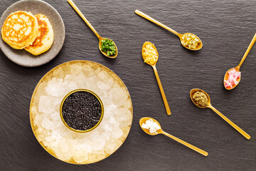 High quality real black sturgeon caviar in golden box on crushed ice aside blinis in a small plate and golden spoons with pickles and herbs on a slate background. Flat Lay. Top view.