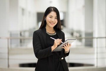 Portrait of a young cheerful businesswoman surfing social network on digital tablet in front of office during break. Asian business woman standing in office building. Business stock photo