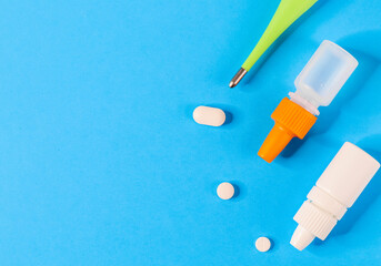 Thermometer, pills and two bottles with drops for nose and eyes on the right against a blue background and with a place for text on the left, top view close-up.