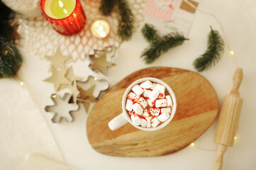 mug of hot cocoa with marshmallows and sprinkles, wooden cutting Board, a small rolling pin, tree branches, burning candles and metal cookie cutters on a beige light Christmas table