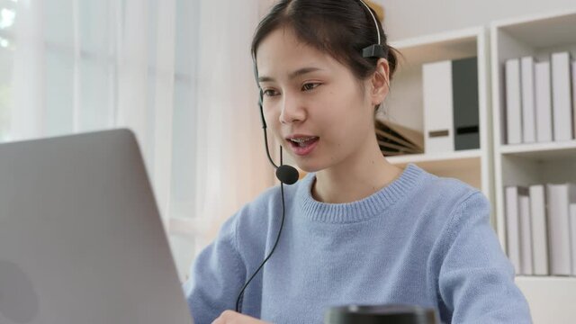 Smiling Asian business woman in casual clothes and wearing microphone headset of customer support phone operator at workplace.	
