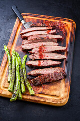 Traditional barbecue sliced dry aged wagyu flank steak offered with green asparagus and chili powder as close-up on a rustic burnt wooden cutting board