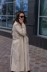 Naklejka premium young brown-haired beautiful girl with green eyes and a light trench with a backdrop of buildings and commercial centers in the city