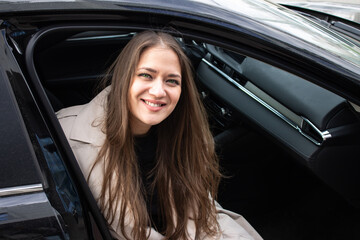 Fototapeta na wymiar beautiful young girl with brown hair green eyes in the bright trench sits in a black car