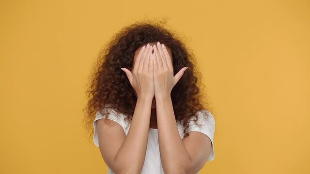 Young african woman girl 20s years old in white t-shirt posing isolated on yellow background studio. People lifestyle concept. Look out camera charming smile cover face with hands play hide and seek