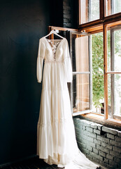 Modern wedding dress hanging at window in soft morning light. Stylish luxury  wedding dress with lace floral pattern in light.