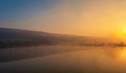 Sunrise over foggy lake. Canes on foreground on the lake coast. Sun is rising up over the trees on the further river bank.