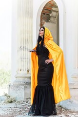 Naklejka premium Mystical woman in yellow cloak, black dress, and tarot cards at old town, Halloween date, party outfit 