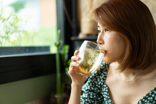 A Young Asian Woman With Dark Brown Hair Wearing A Green Floral Pattern Sipping Iced Tea In A Clear Glass By The Window Of A Coffee Shop.