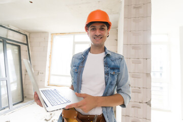 Handsome architect or foreman in helmet working with laptop on the apartment drawings at the structure white interior