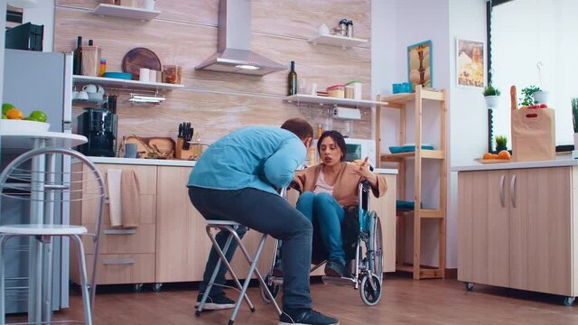 Unhappy disabled wife in wheelchair because of disagreement with husband in kitchen. Woman with paralysis handicap disability handicapped difficulties getting help for mobility from love and