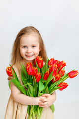 Little beautiful girl blonde with a bouquet of tulips on a light background. Baby girl smiling. Spring and women's day concept. Little girl holding a bouquet of red tulips.