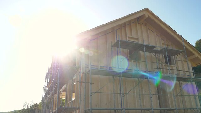CLOSE UP, LENS FLARE: Bright spring sunbeams shine on a large wooden house being built in countryside. Scenic view of an unfinished cross-laminated timber house being built in rural area on sunny day.