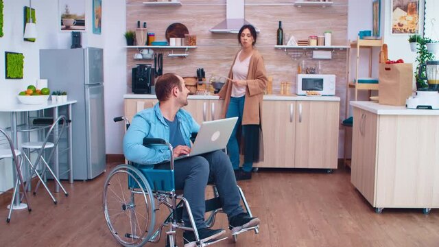 Handicapped entrepreneur in wheelchair working on laptop while wife is preparing meal in kitchen. Man with paralysis handicap disability handicapped difficulties getting help for mobility from love