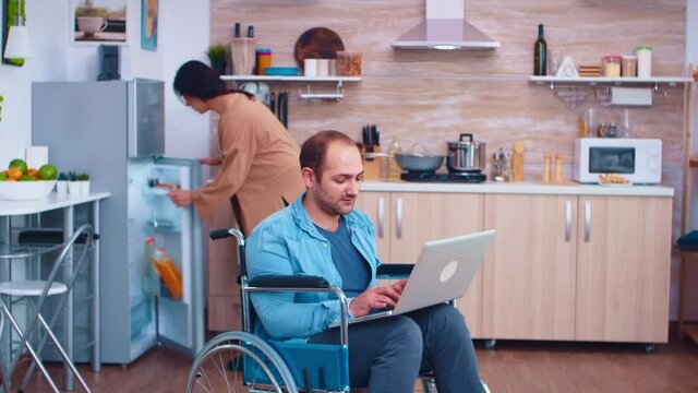 Disabled businessman in wheelchair working on laptop while wife is cooking in kitchen. Corporate man with paralysis handicap disability handicapped difficulties working after accident having internet