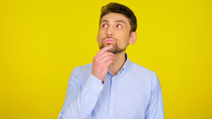 Handsome bearded man looks away on yellow background with copy space. Guy in light blue shirt with hand on beard looks in place for text or goods