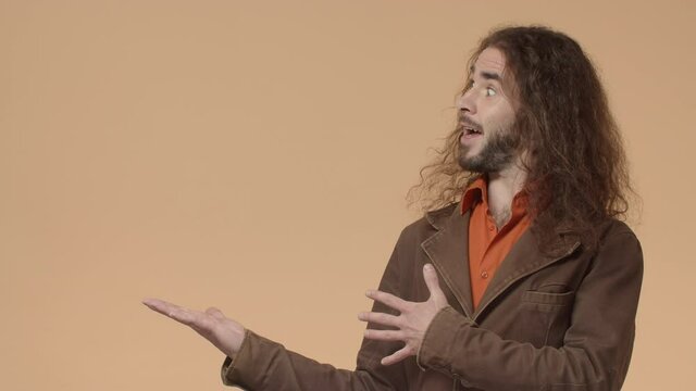 Handsome bearded hipster guy with long curly hair say ta-da, fanfare to introduce awesome product, pointing hands left, presenting item over beige background, make announcement
