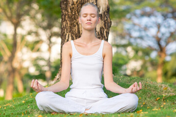 Beautiful relaxed woman meditating while sitting on the grass under a tree at the park. Spiritual yoga and healthy calmness. 