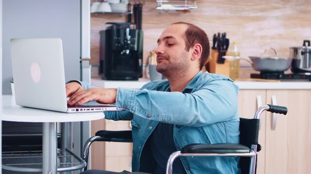 Man with special needs in wheelchair working on laptop in kitchen. Corporate man with paralysis handicap disability handicapped difficulties working after accident having internet online video call