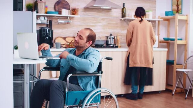 Tired disabled man in wheelchair working on laptop in kitchen and wife is cooking meal. Corporate man with paralysis handicap disability handicapped difficulties working after accident having internet