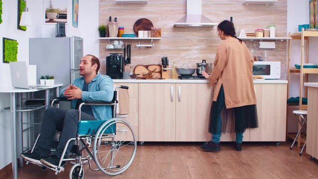 Happy disabled man in wheelchair using laptop in kitchen. Man talking on video conference. Corporate man with paralysis handicap disability handicapped difficulties working after accident having