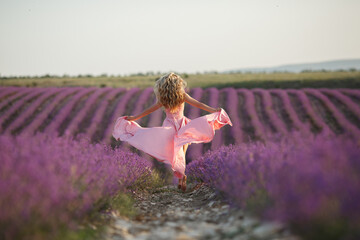 pretty blonde woman running away in lavender field. woman in long dress and straw hat having fun in flowers of lavender