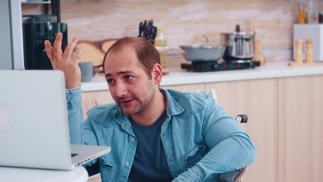 Handicapped businessman talking during video call on laptop in kitchen. Woman cooking. Corporate man with paralysis handicap disability handicapped difficulties working after accident having internet