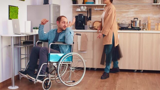 Smiling disabled man in wheelchair and his wife during a video call on laptop in kitchen. Corporate man with paralysis handicap disability handicapped difficulties working after accident having