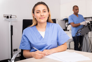 Positive female doctor cosmetologist wearing blue overall filling out medical forms with male assistant preparing equipment for procedures on background