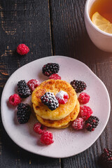 Cottage cheese pancakes, curd fritters dessert with raspberry and blackberry berries in plate near to hot tea cup with lemon slice on dark black wooden background, angle view