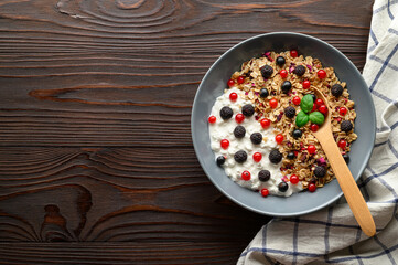 Healthy breakfast. Bowl with oat granola, yogurt and black raspberries, black and red currants on a brown wooden background. Top view, copy space.