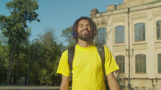 Joyful happy stylish african american male college student in headphones enjoying music, swaying to the beat, expressing positive vibes and carefree mood while walking on university campus to lessons